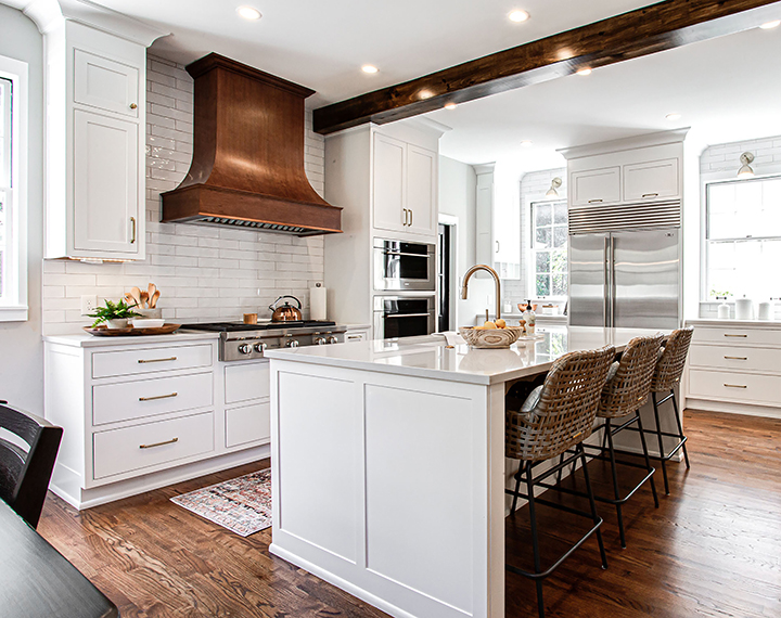 White semi-custom kitchen cabinets, natural wood floors, with a white island. There is a beam in the center of the ceiling that matches the floor and a stained hood above the stove top.