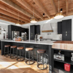 A large kitchen that has white subway tiled walls. The cabinets in the kitchen are black. The countertops are white except for one area in the kitchen that has a butcher block countertop over some additional storage. This butcher block has a joint to overlap onto the white counter top of the larger island for a seamless look. The roof of the kitchen has exposed beams with round gold light fixtures.