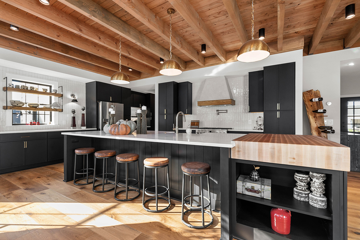 A large kitchen that has white subway tiled walls. The cabinets in the kitchen are black. The countertops are white except for one area in the kitchen that has a butcher block countertop over some additional storage. This butcher block has a joint to overlap onto the white counter top of the larger island for a seamless look. The roof of the kitchen has exposed beams with round gold light fixtures.