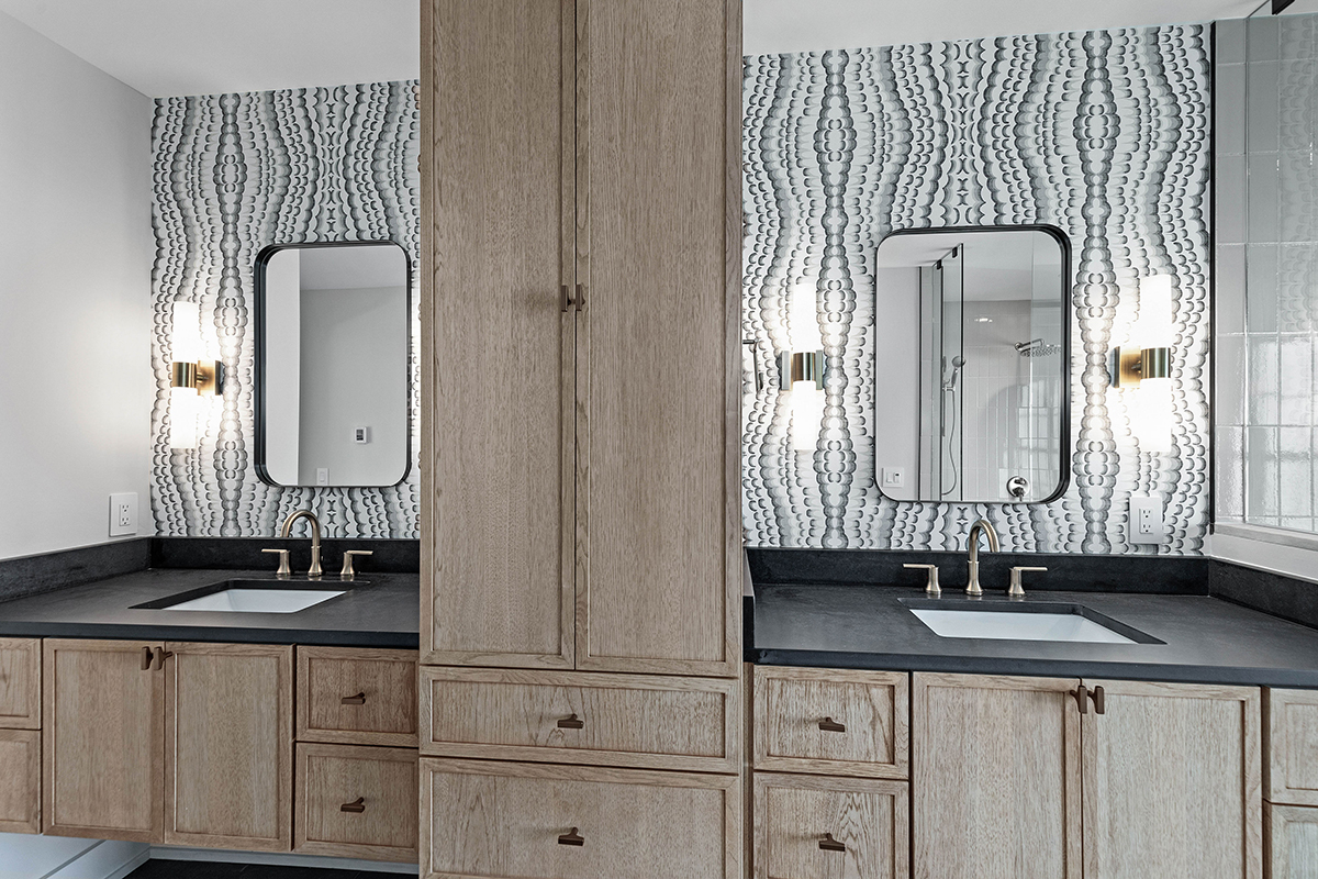 shows dual sinks with natural grey wood cabinets. behind the black counters there is snake skin wallpaper. there is a large floor to ceiling linen cabinet between sinks.