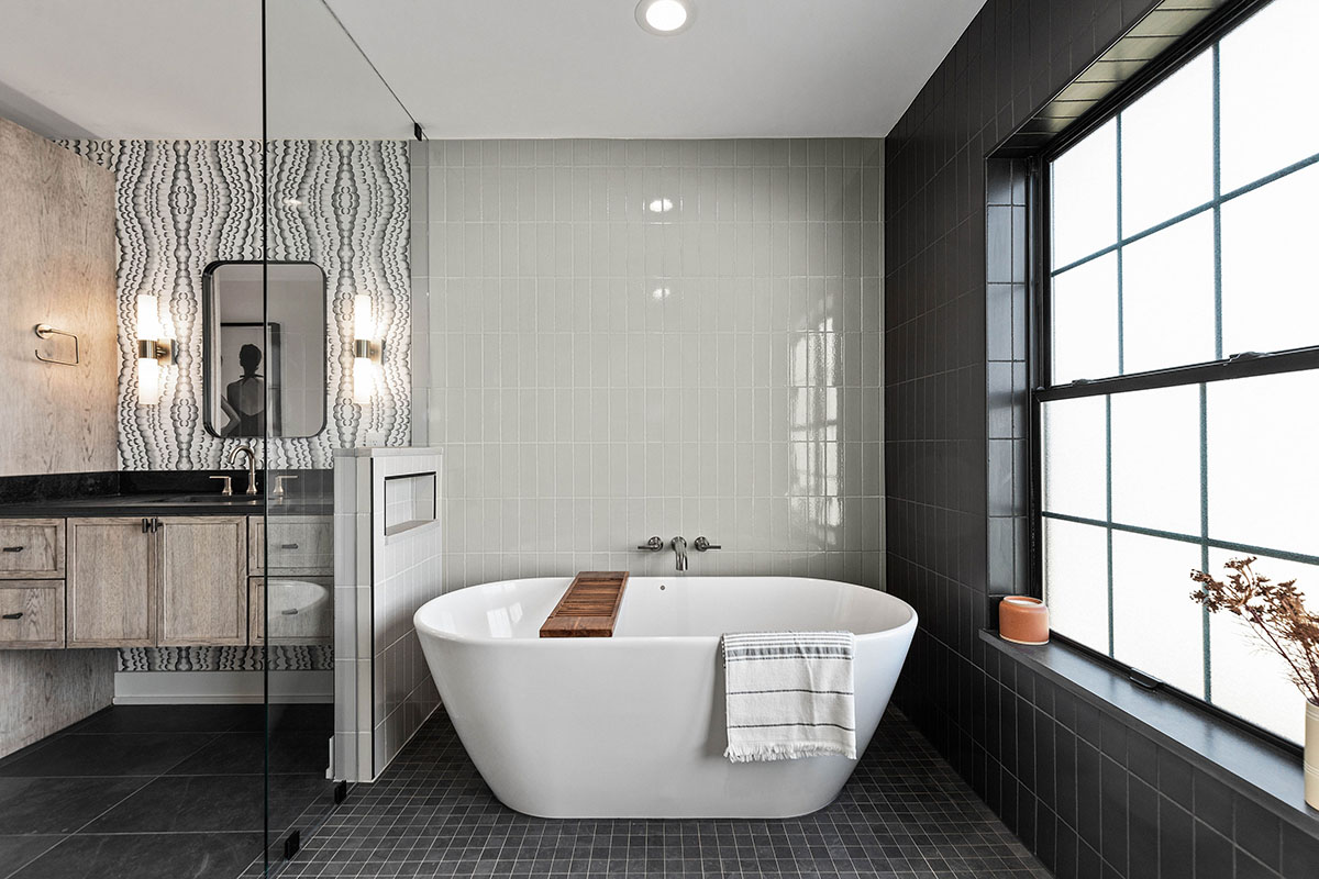 black and white bathroom with large white soaking tub. the cabinets are light grey but look like natural wood. the bathroom counter is dark black. there are several different tiles around this bathroom to help separate the bathroom into different areas.