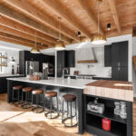 black and white kitchen with black cabinetry and white stone countertops. Large cutting board area on the edge of the island. Brown wood beamed ceilings hang overhead the brass lights.