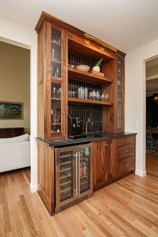 Brown wood minibar with glossy black stone backsplash. There is glassware in the storage compartments and drinks in the wine fridge.