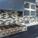 Multicolored tile backsplash accompanied with stainless steel appliances and glossy blue kitchen cabinetry.