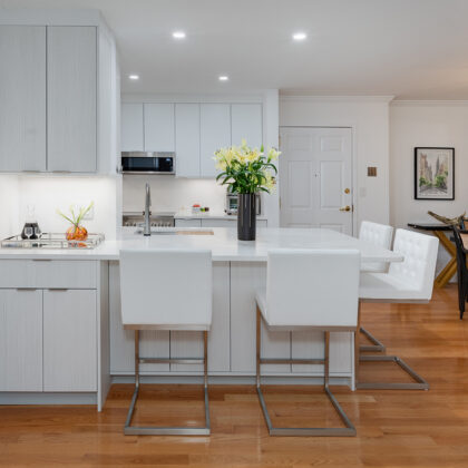 Ultra white kitchen with white cabinets and white stone counter tops. the dining room is also shown but is set apart from the kitchen as it has dark brown table with a dark-brushed lighting fixture overhear. the floor is a natural wood.