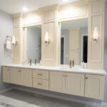 Light blue bathroom with cream double vanity. The cabinets are floating off of the floor but there is molding around the mirror that goes all the way up to the ceiling. The fixtures in the bathroom are gold