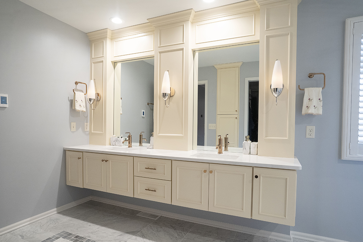 Light blue bathroom with cream double vanity. The cabinets are floating off of the floor but there is molding around the mirror that goes all the way up to the ceiling. The fixtures in the bathroom are gold
