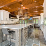 ski house with stone flooring, wooden ceilings and cabinetry. The stone countertop sits on faded wood, while the kitchen cabinetry is all white. The lights above the ceiling are modern fixtures. White brick backsplash aligns the walls.