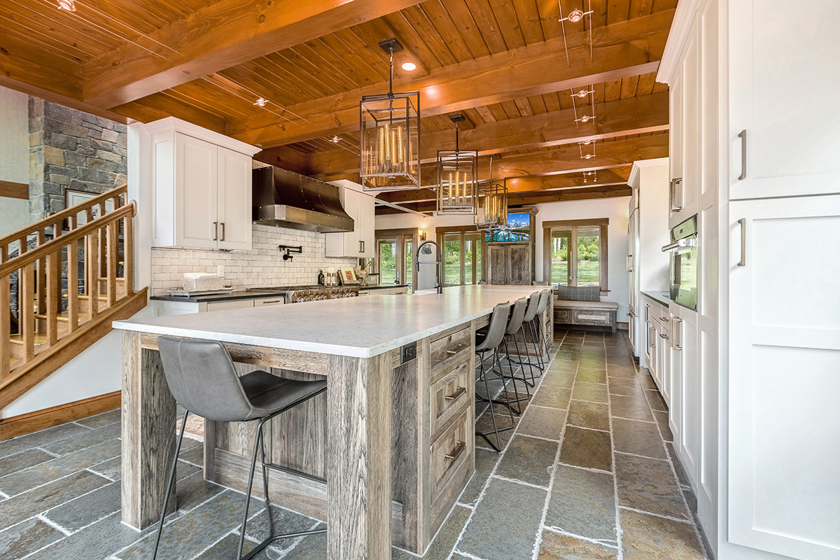ski house with stone flooring, wooden ceilings and cabinetry. The stone countertop sits on faded wood, while the kitchen cabinetry is all white. The lights above the ceiling are modern fixtures. White brick backsplash aligns the walls.