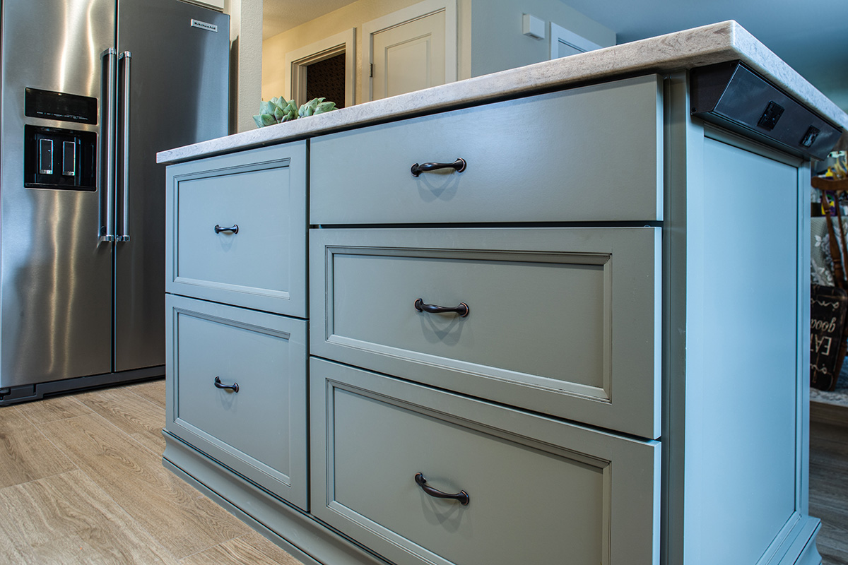 A view of the kitchen island. There are 3 doors stacked on the right half of the island. The left half of the island has two larger drawers. The island is a soft green color with dark brass handles. We can also see under counter plug-ins that have been installed on the island.
