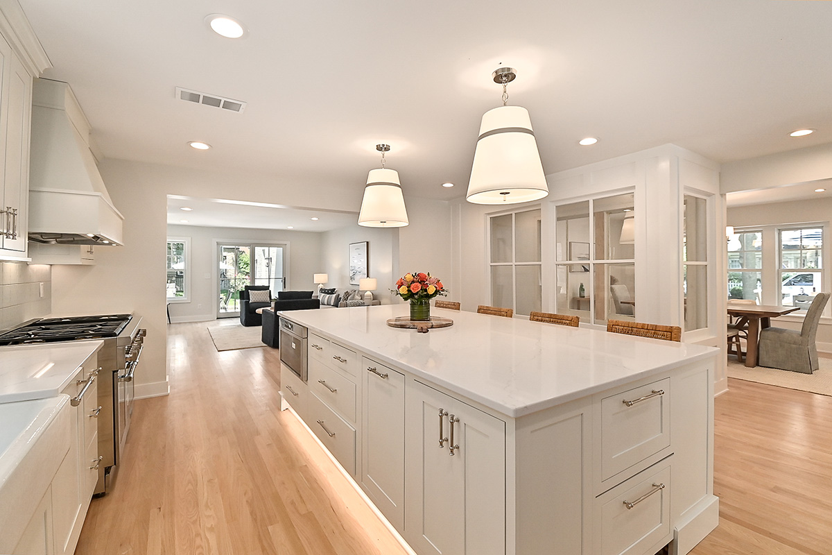 all white kitchen with silver accents. Well lit open floorplan extending to seating area and dining room. White countertops, and white wooden cabinets.