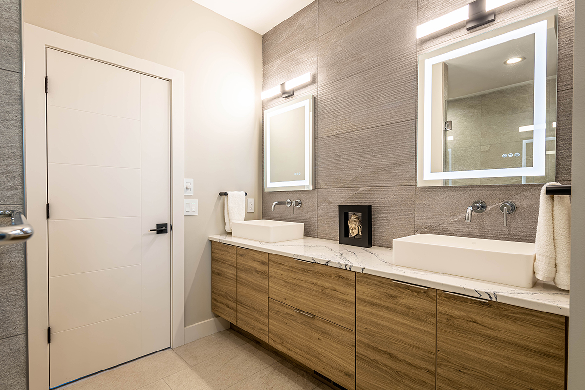 home bathroom, with modern wood cabinets that have sleek hidden handles. There are tech ingrained mirrors and above-counter sinks on a white granite counter top.