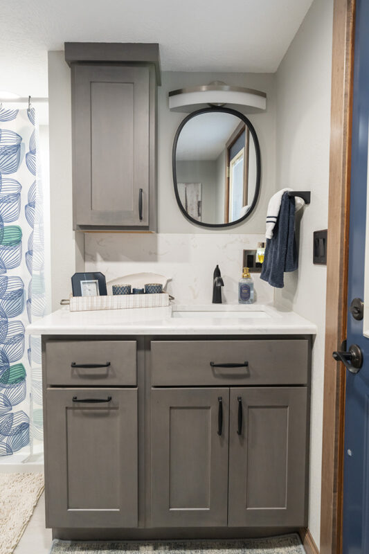 This photo is a bathroom that has an outdoor entry. The cabinets under the sink are slate grey and have black handles. The counter is white. All hardware of this bathroom is black.