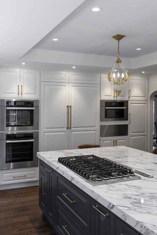 Dual tone kitchen is pictured. The island has black cabinets with a marbled counter top and the flat top is built into the island. In the distance, there is a wall that has stoves, a microwave oven, and large pantry. The cabinets surround these appliances are white with large gold handles.