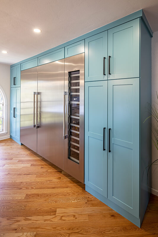 large stainless steel fridge surrounded by turquoise blue cabinets on bright wooden floor. The handles of the turquoise cabinets are black and sleek.