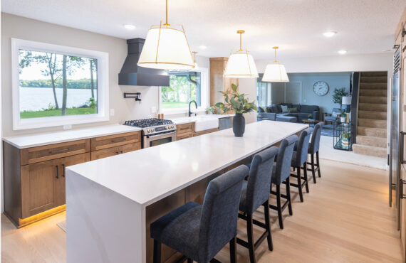 Open kitchen with long island with seating facing stained base cabinets, oven, hood, sink and large windows overlooking a lake.