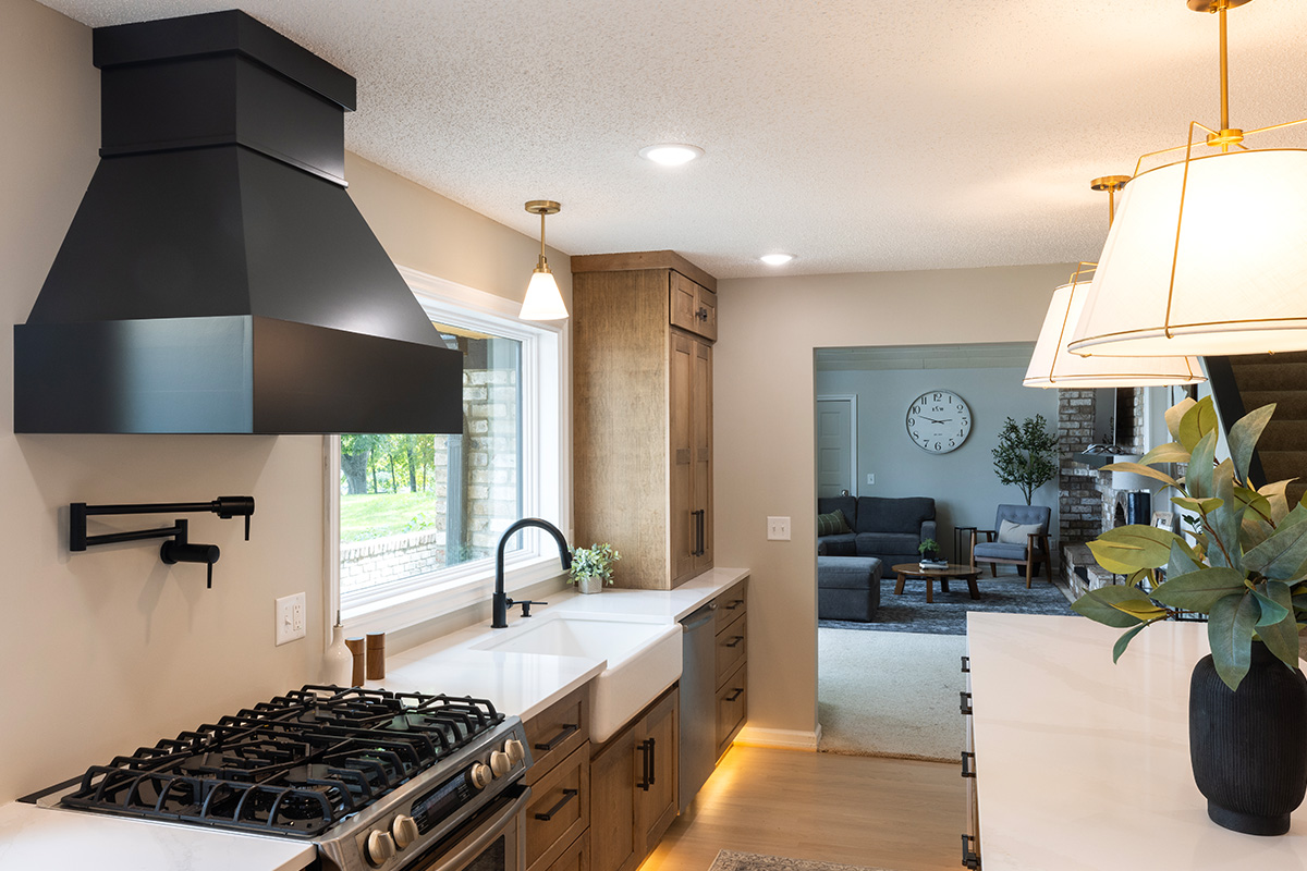 Open kitchen with long island with seating facing stained base cabinets, oven, hood, sink and large windows overlooking a lake.