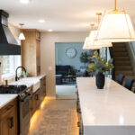 Open kitchen with long island with seating facing stained base cabinets, oven, hood, sink and large windows overlooking a lake.