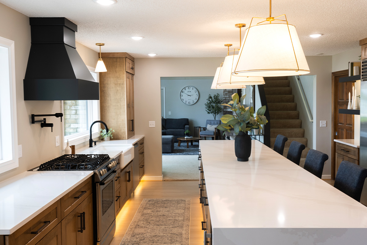 Open kitchen with long island with seating facing stained base cabinets, oven, hood, sink and large windows overlooking a lake.