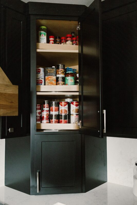 Black painted kitchen cabinets. Showing open door of a countertop cabinet with food items inside.