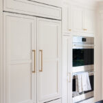 White kitchen. View of fridge covered with decorative panels and stacked ovens.