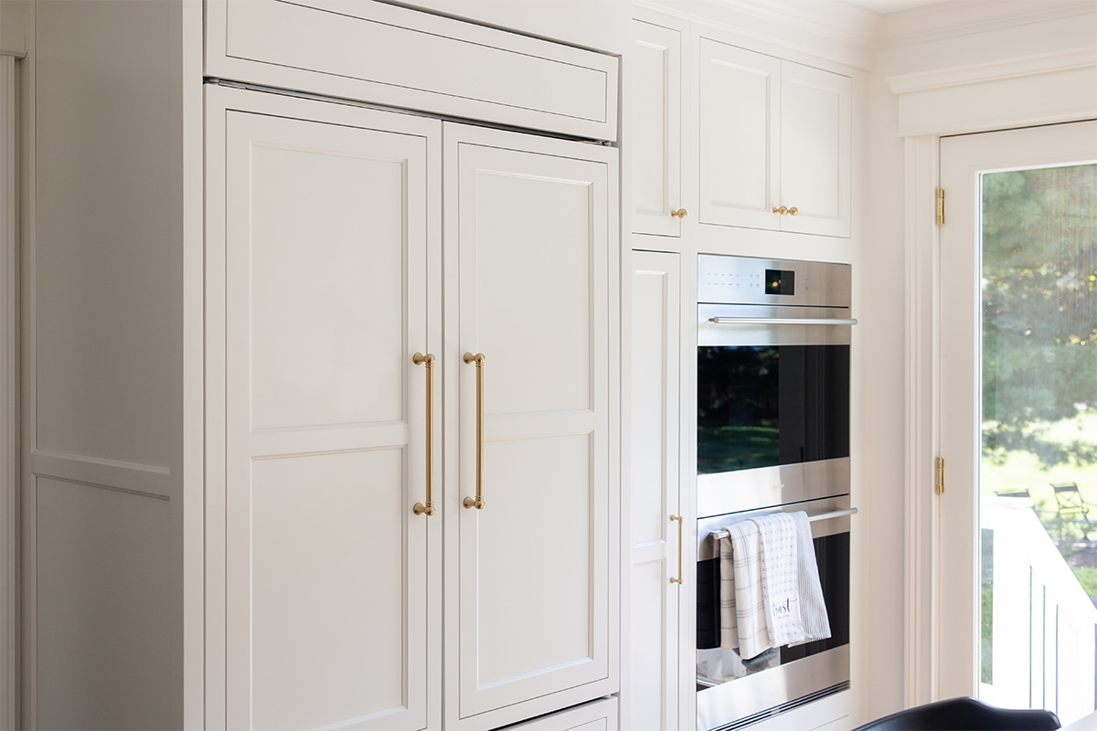 White kitchen. View of fridge covered with decorative panels and stacked ovens.