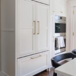 White kitchen. View of fridge covered with decorative panels and stacked ovens.