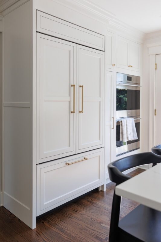 White kitchen. View of fridge covered with decorative panels and stacked ovens.