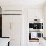 White kitchen. View of fridge covered with decorative panels and stacked ovens.