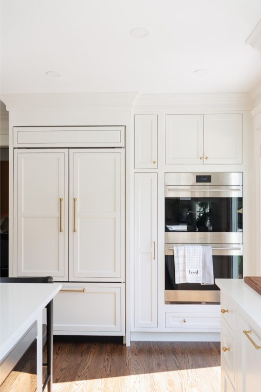 White kitchen. View of fridge covered with decorative panels and stacked ovens.