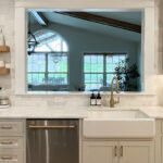 White painted kitchen looking through a window out to a great room.