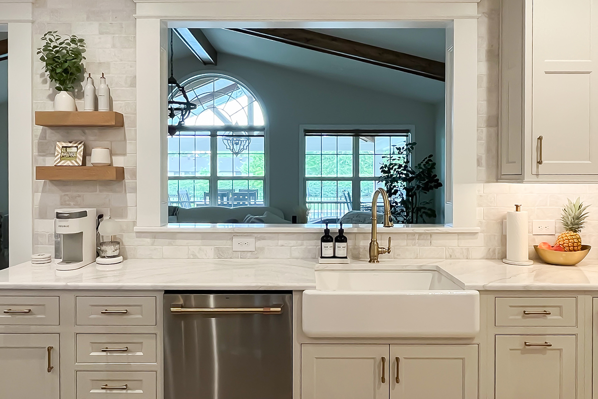 White painted kitchen looking through a window out to a great room.