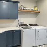 Blue painted laundry room cabinets with white appliances and stained floating shelves