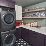 Purple stacked washer and dryer next to purple cabinets with stained wood floating shelves.