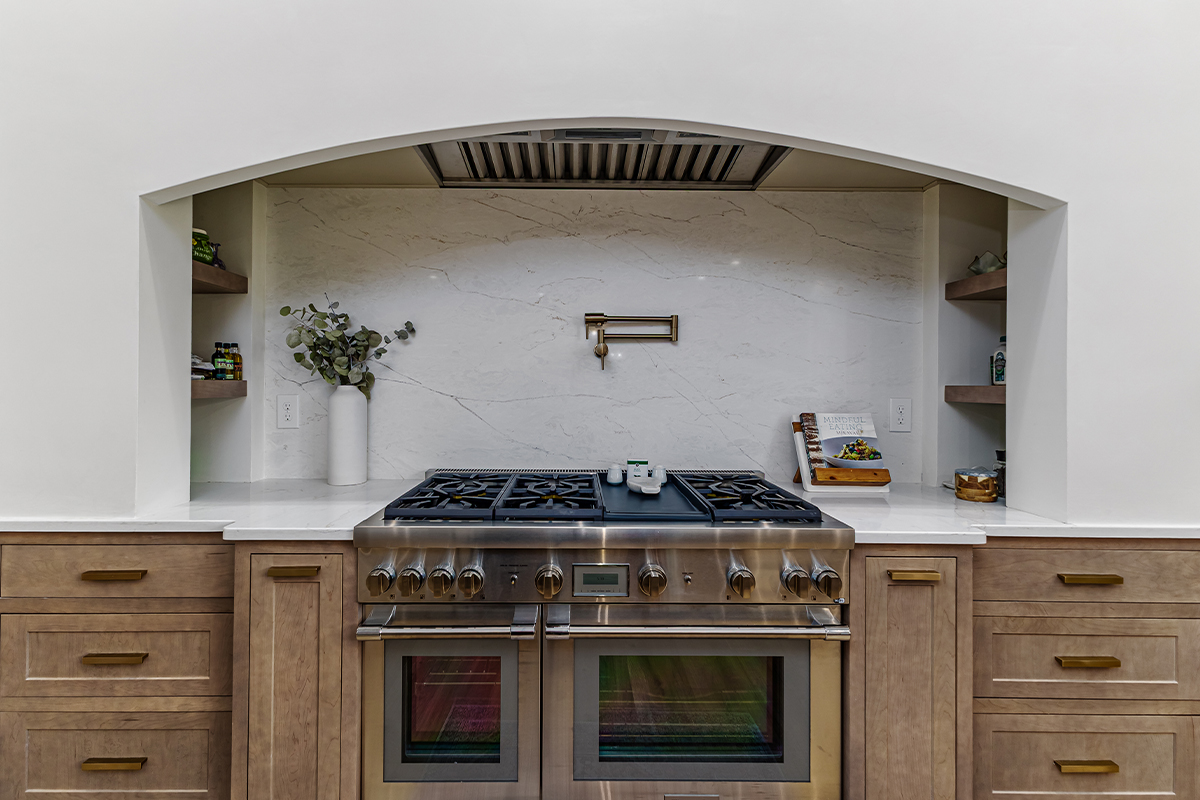 Light brown-stained lower and kitchen cabinets with gray painted archway blower above a professional range.