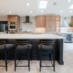 Open kitchen with perimeter cabinets in Sable stain on rift white oak and a dark island in Midnight stain.
