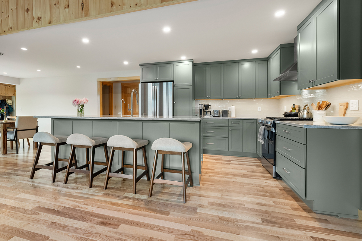 Green kitchen cabinets with island seating and light brown wood floors.