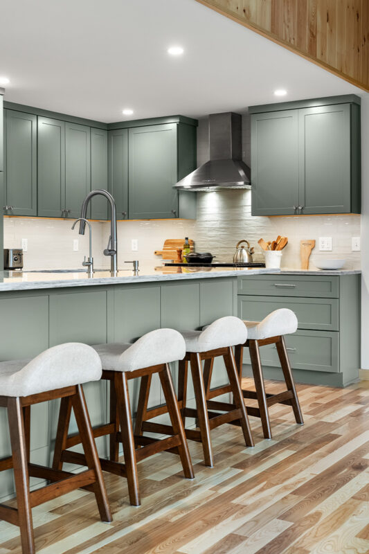 Green kitchen cabinets with island seating and light brown wood floors.