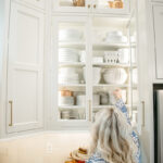 Woman reach up to a white kitchen cabinet with glass doors.