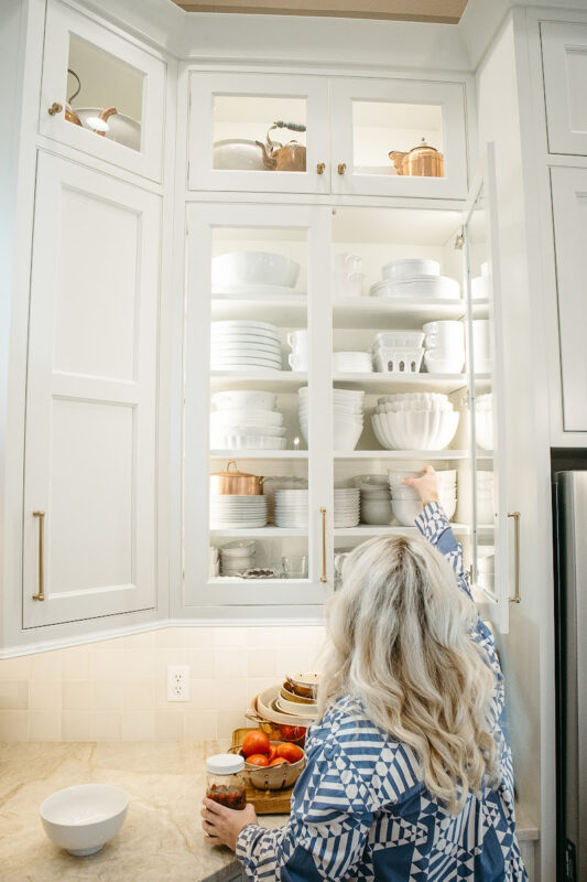 Woman reach up to a white kitchen cabinet with glass doors.